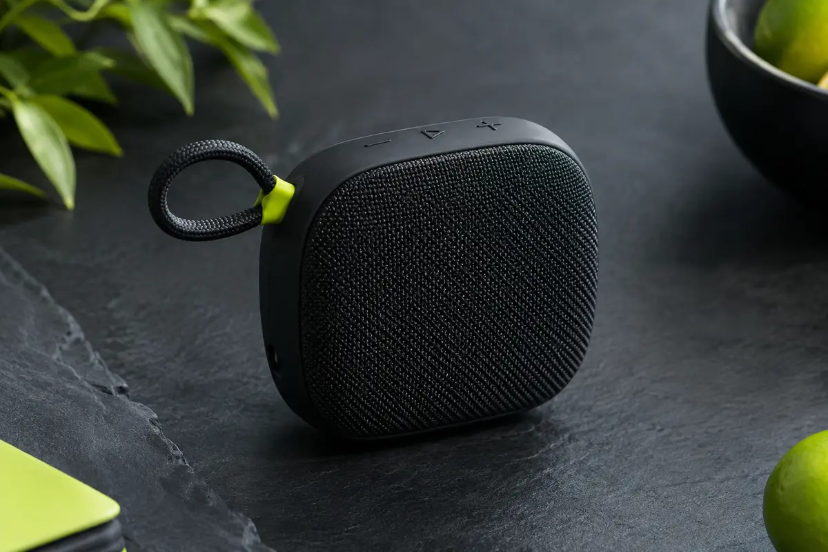Enceinte Bluetooth noire compacte avec grille textile et boucle intégrée, présentée sur fond ardoise